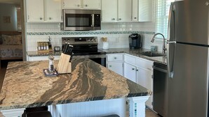 A kitchen with white cabinets, a granite island with bar stools, a stainless steel refrigerator, and a microwave.