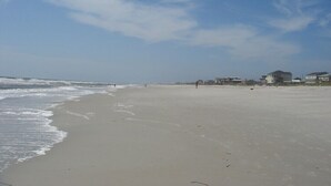 A wide sandy beach with gentle waves, a clear sky, and a few buildings in the distance.