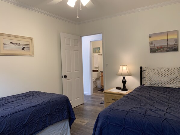 A bedroom with two beds, a nightstand with a lamp, a painting on the wall, and a door leading to another room. - 2 BR on Jersey Ct Just Steps to The Beach (San Diego)
