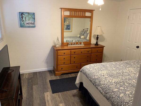 Quarto com penteadeira de madeira, cama com colcha florida, mesa de cabeceira, abajur, espelho e um pequeno objeto decorativo na penteadeira.