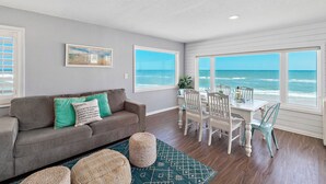 A living room with a gray sofa, a dining table with white chairs, and large windows overlooking the ocean.