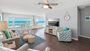 A spacious living room with a large window overlooking the ocean, a ceiling fan, a TV, and a cozy seating area.