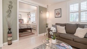 A modern living room with a sofa, coffee table, and decorative items. A hallway leads to a bedroom with a bed and a dresser. - Beautiful 1 Bedroom Apartment minutes to SFO in Victorian Home (Can sleep 4) (South San Francisco)