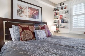 Une chambre bien rangée, avec un grand lit, une bibliothèque et des œuvres d’art encadrées accrochées au mur.