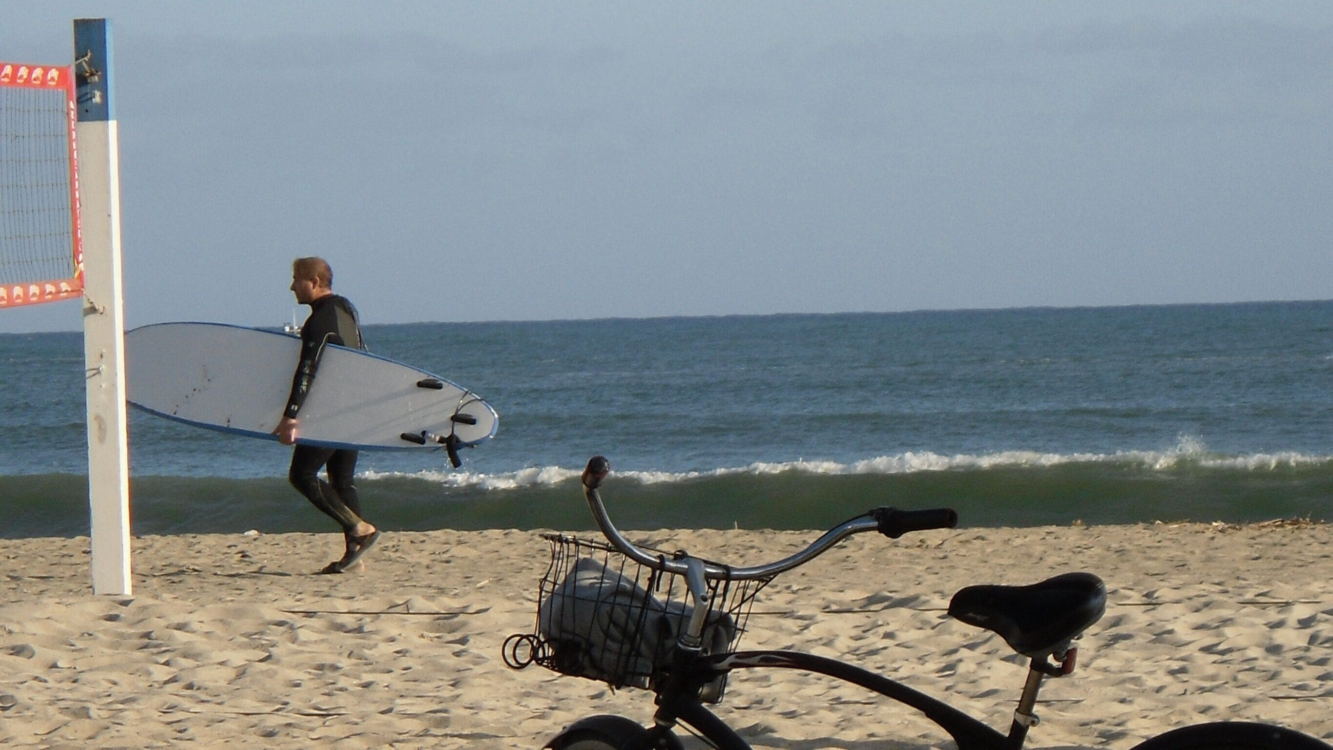 A beach scene with a person carrying a surfboard and a bicycle with a basket.