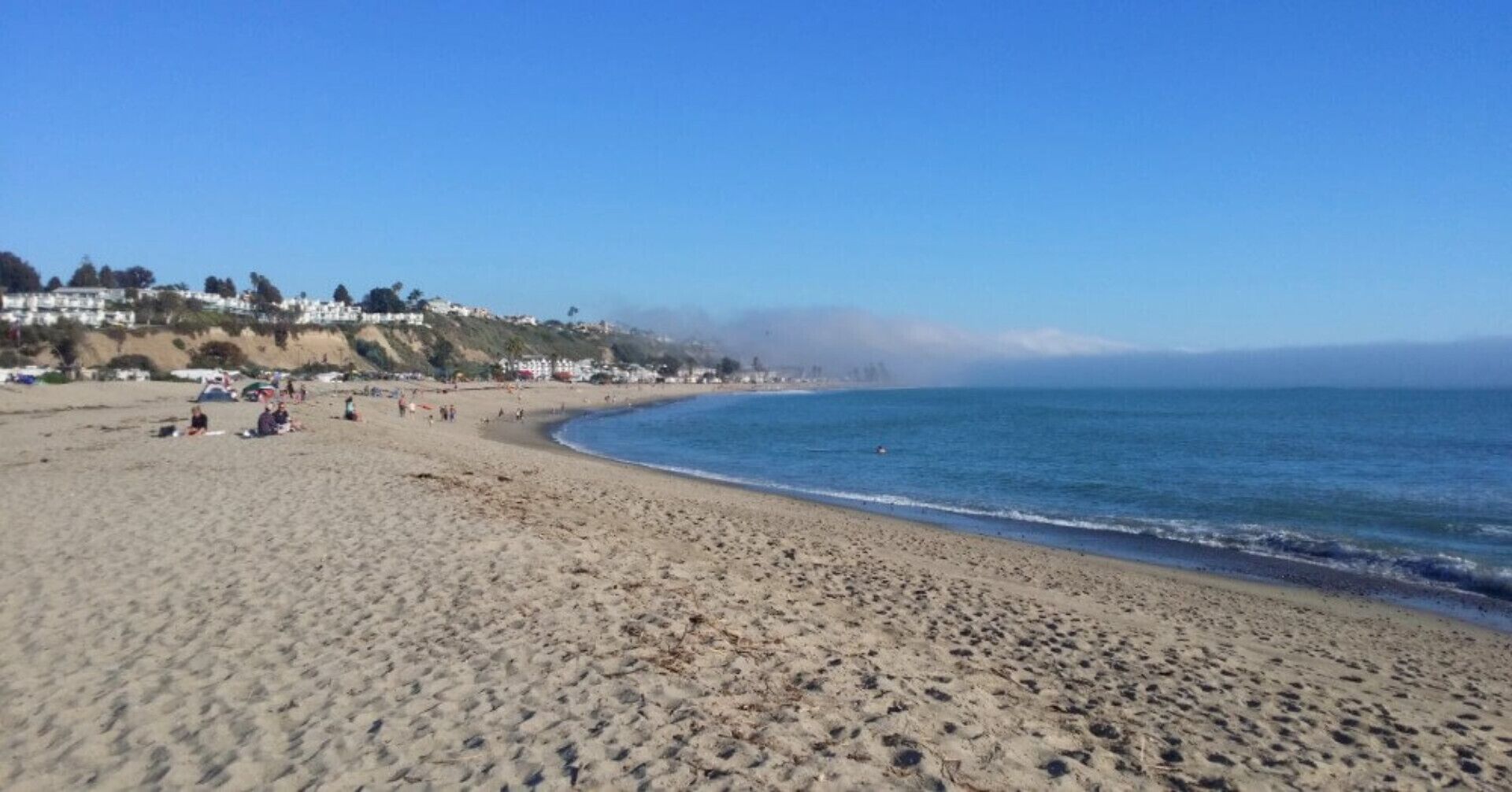 Una extensa playa arenosa con un océano azul cristalino, algunas personas y casas en una colina al fondo.