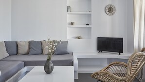 Una sala moderna con un sofá gris, una mesa de centro blanca con un florero, un televisor sobre un mueble blanco y una silla de mimbre.