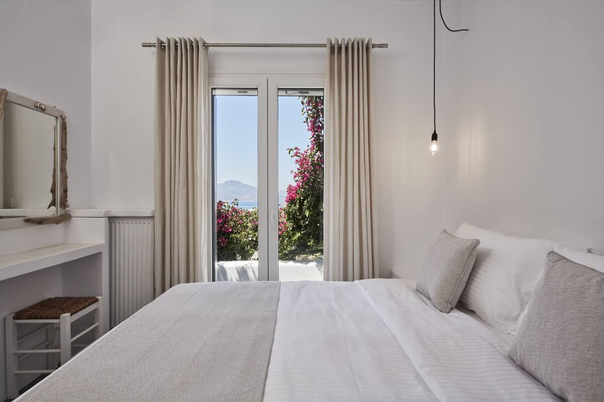 A bedroom with a large bed, a view of pink flowers through the window, and a white dresser.