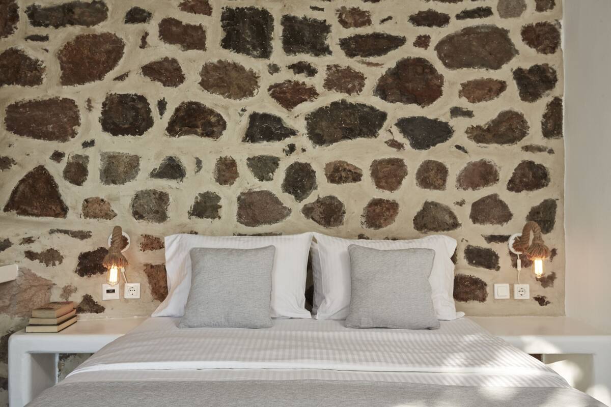 A bed with white bedding and two pillows, a stone wall behind it, and two wall-mounted light fixtures.