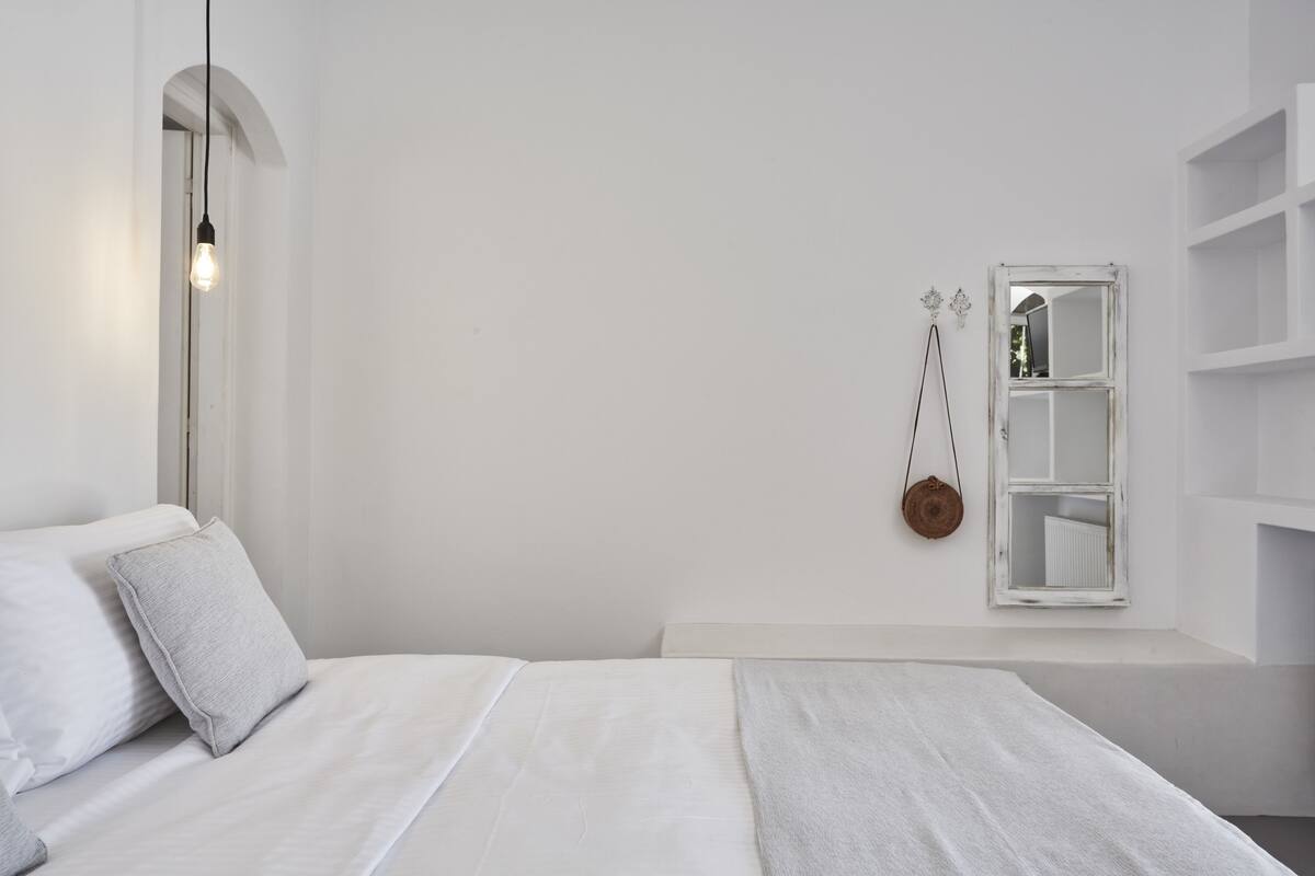 A minimalist bedroom with a bed, a hanging light, a mirror, and a shelf unit.
