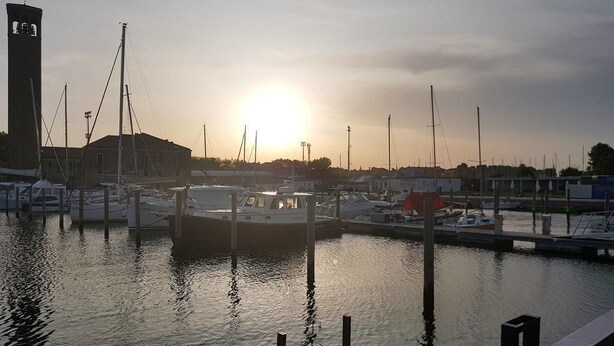 A marina with boats docked, a building with a tower, and a sunset in the background. - Yacht Diva (Venice)