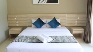A neatly made bed with a patterned bedspread, two pillows, and a rolled towel. There are two bedside tables with lamps, a wall-mounted artwork, and a curtain on the right. - Rusty Seminyak (Seminyak)
