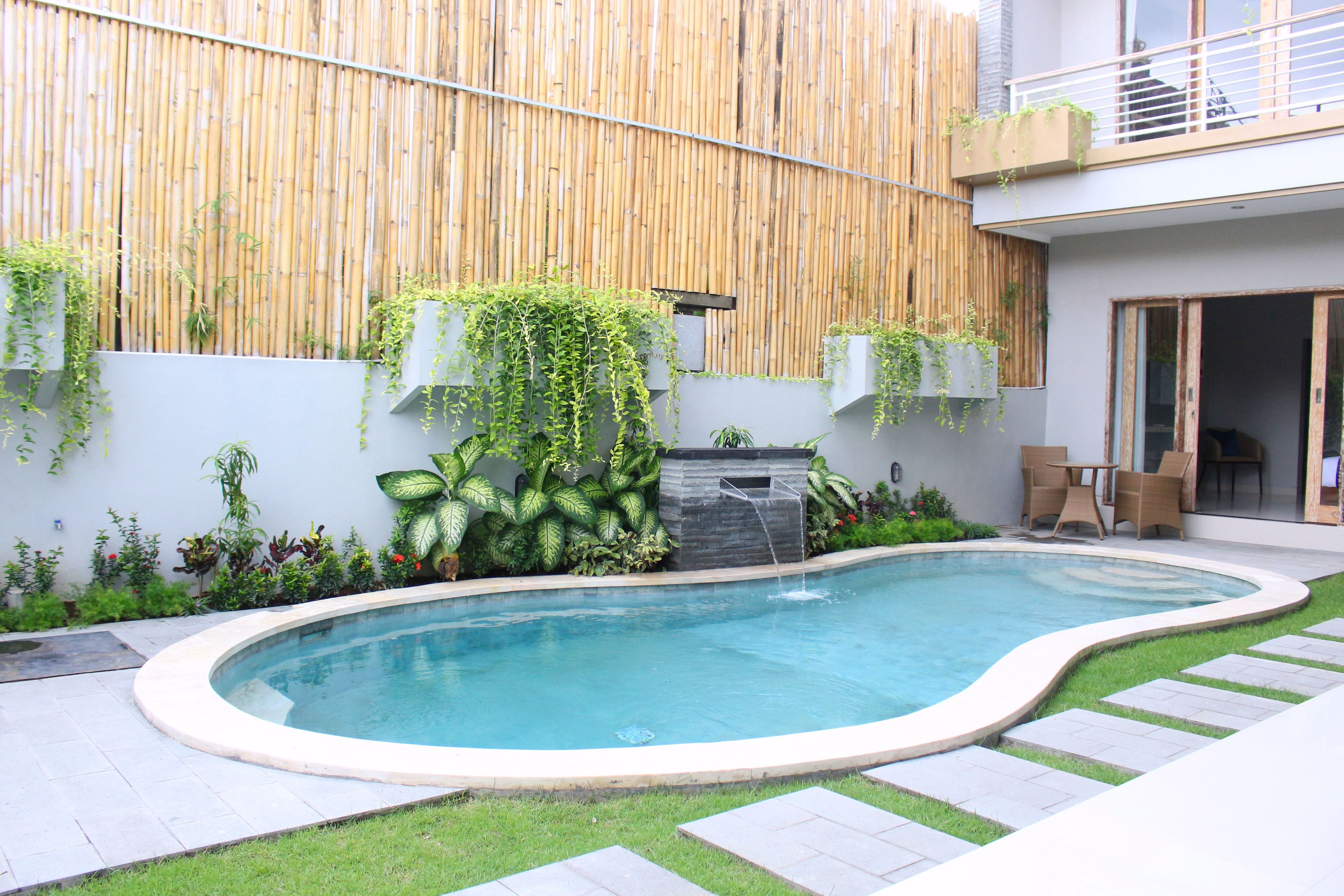 A modern outdoor pool area with a bamboo wall, a small waterfall feature, and a seating area with a table and chairs.