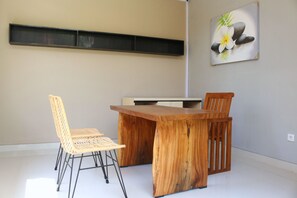 A dining area with a wooden table and two chairs, a wall-mounted shelf, and a painting of flowers and rocks.