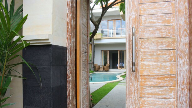 A view through an open wooden door into a courtyard with a swimming pool, a small garden, and a building with a balcony. - Rusty Seminyak (Seminyak)