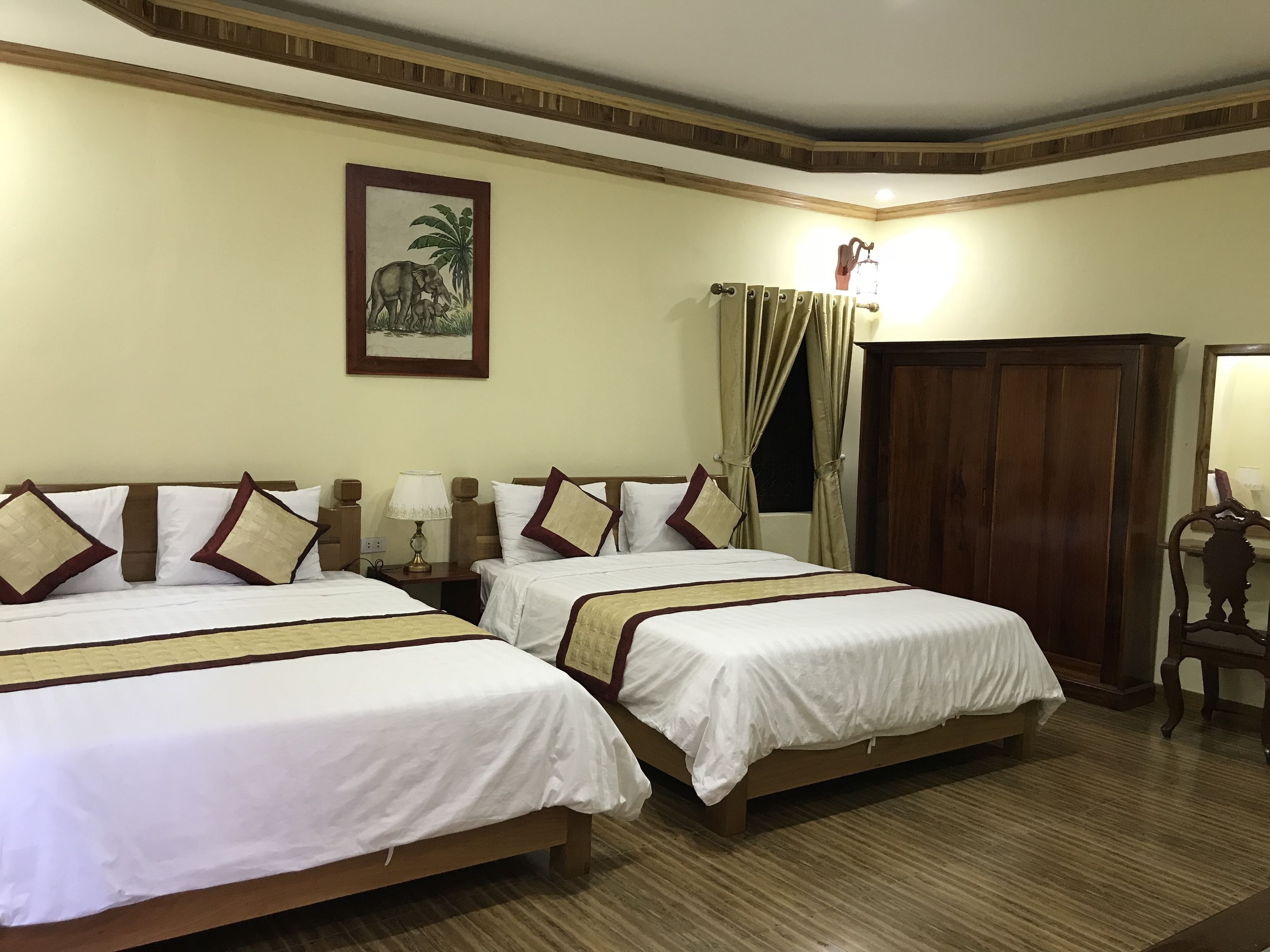 A hotel room with two beds, a wooden wardrobe, a nightstand with a lamp, and a painting on the wall.