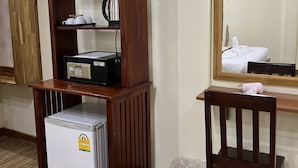 Ein kleiner, hölzerner Schrank mit integriertem Minikühlschrank, Wasserkocher und Kaffeemaschine. Ein Stuhl steht daneben, und ein Mülleimer ist auf dem Boden sichtbar.