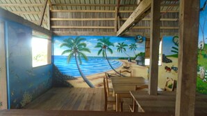 A room with a beach mural, wooden furniture, and a high ceiling. - Emeraude lodge (Antsiranana)