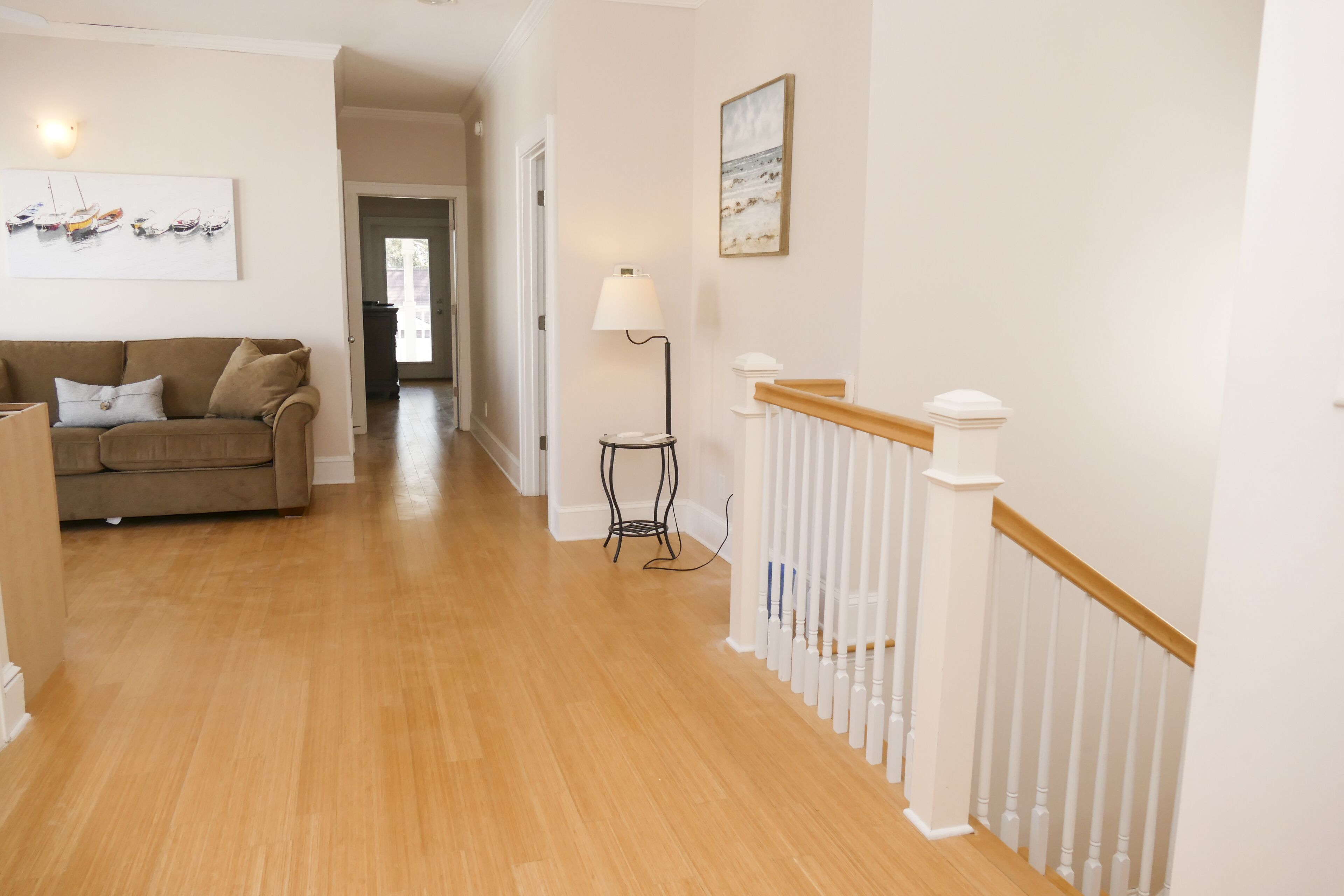 Un couloir doté d’un plancher en bois, d’un canapé beige, d’une lampe et d’un escalier avec des rampes blanches.