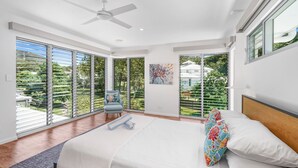 A modern bedroom with a large bed, a ceiling fan, and a view of greenery through floor-to-ceiling windows. - Belle Escapes Tranquil Waters Luxury Home Palm Cove (Palm Cove)