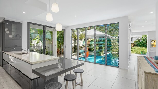 A modern kitchen with a large island, a dining area with stools, and a view of a swimming pool and greenery. - Belle Escapes Tranquil Waters Luxury Home Palm Cove (Palm Cove)