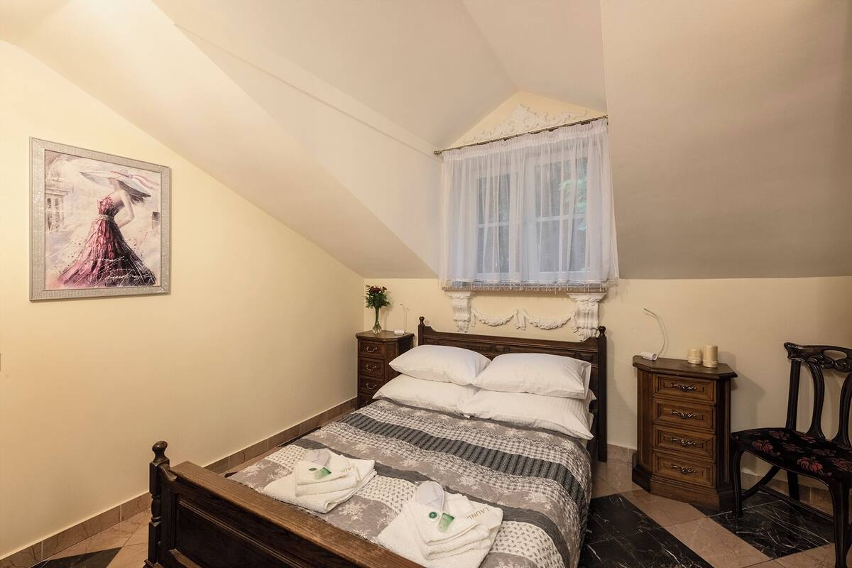 Une chambre à coucher comprenant un lit, une table de chevet, une chaise, une fenêtre avec un rideau léger, un tableau au mur et un vase avec des fleurs.