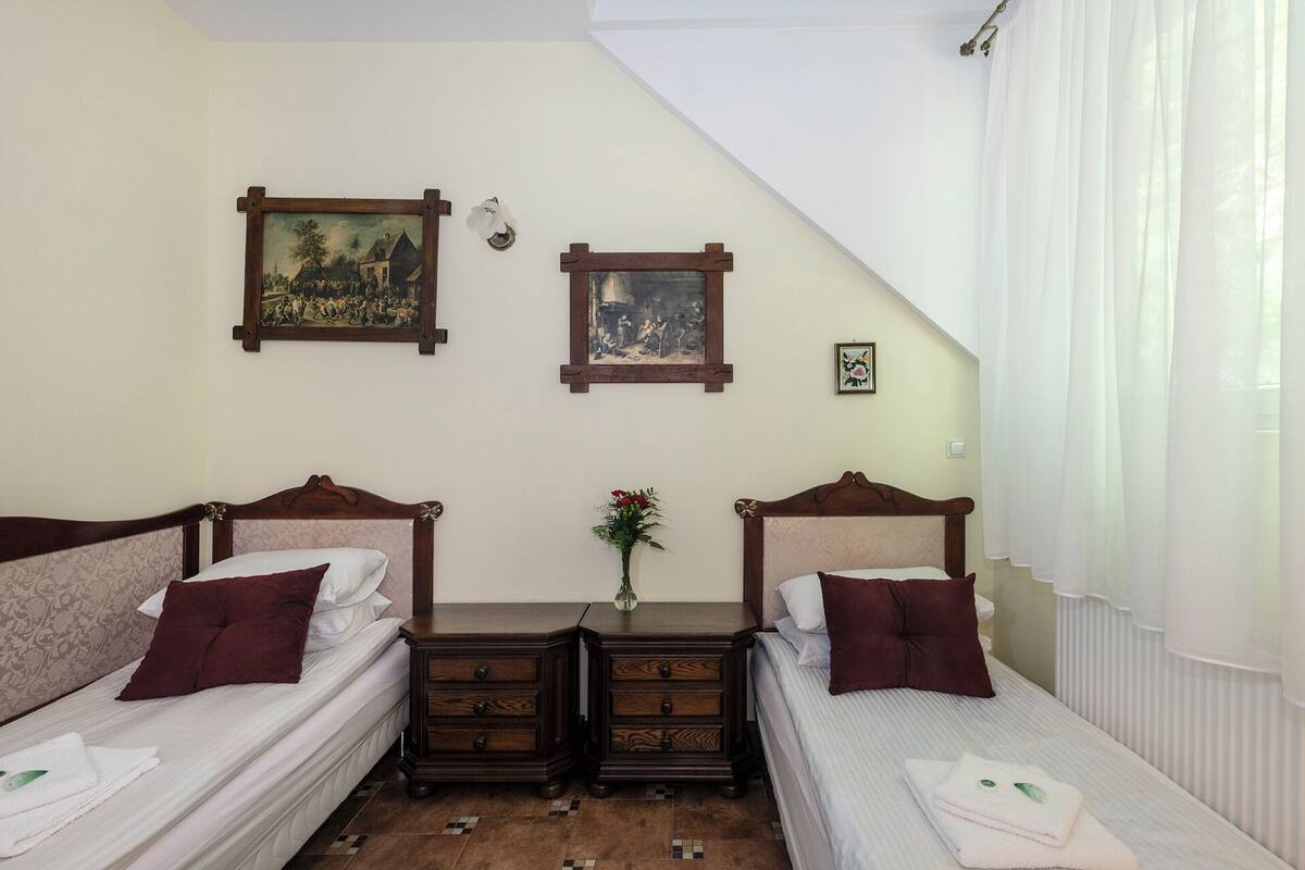 Une chambre avec deux lits simples, une table de chevet avec des tiroirs, une petite table sur laquelle est posé un vase, et deux tableaux encadrés accrochés au mur.