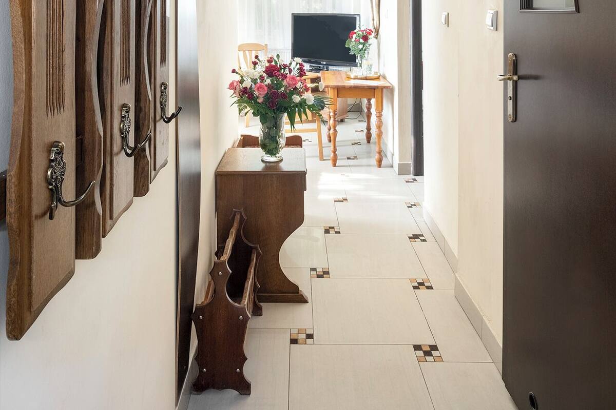 Un couloir avec un porte-manteau en bois fixé au mur, une table en bois sur laquelle est posé un vase de fleurs, un téléviseur et une porte dotée d’un miroir.