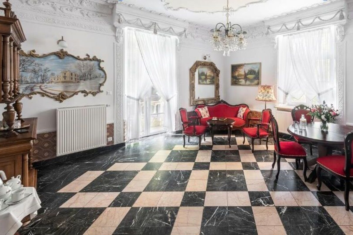 Ein geräumiges Zimmer mit einem Schachbrettmuster-Bodenbelag, einem Esstisch mit Stühlen, einer Couch und einer Chandelier.