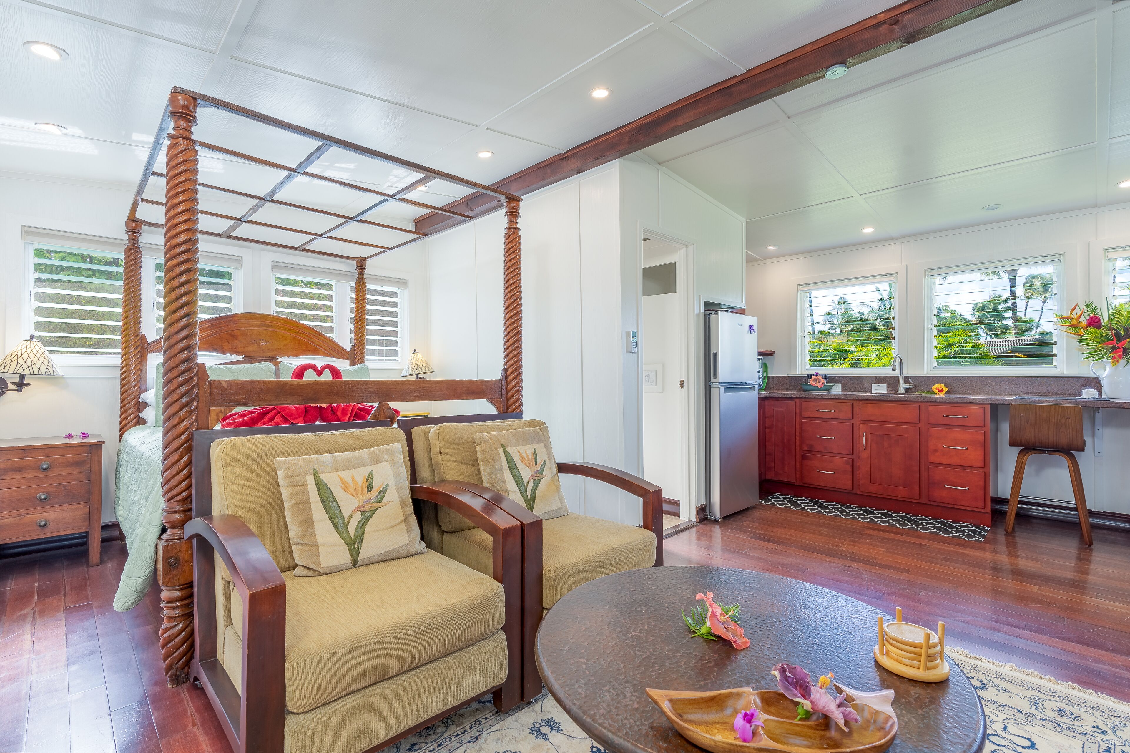A four-poster bed with a wooden frame, a sofa with floral cushions, a round wooden table with decorative items, and a kitchen area with wooden cabinets and a refrigerator.