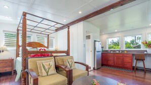 Ein Bett mit Baldachin und Holzrahmen, ein Sofa mit floralen Kissen, ein runder Holztisch mit Dekorationen sowie eine Küchenzeile mit Holzschränken und einem Kühlschrank.
