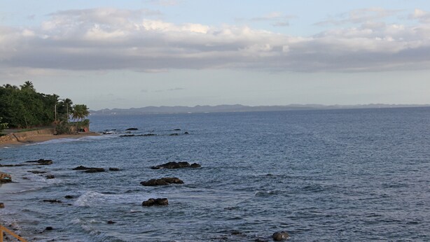 Paisaje costero con playa, costa rocosa y montañas a lo lejos.