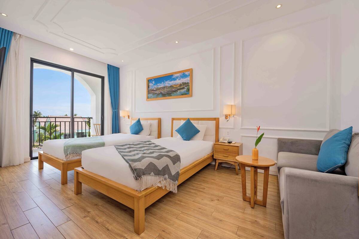 A hotel room with a large bed, a sofa, a small table, and a balcony with curtains.