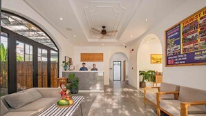 Um saguão de hotel com balcão de recepção, sofás, uma mesa de centro com frutas e um cartaz divulgando passeios turísticos.