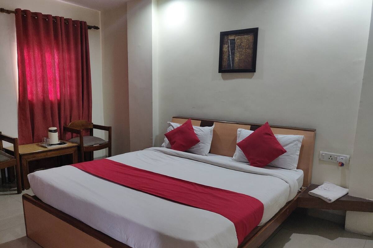 A hotel room with a bed, a desk with a chair, a red curtain, and a framed picture on the wall.