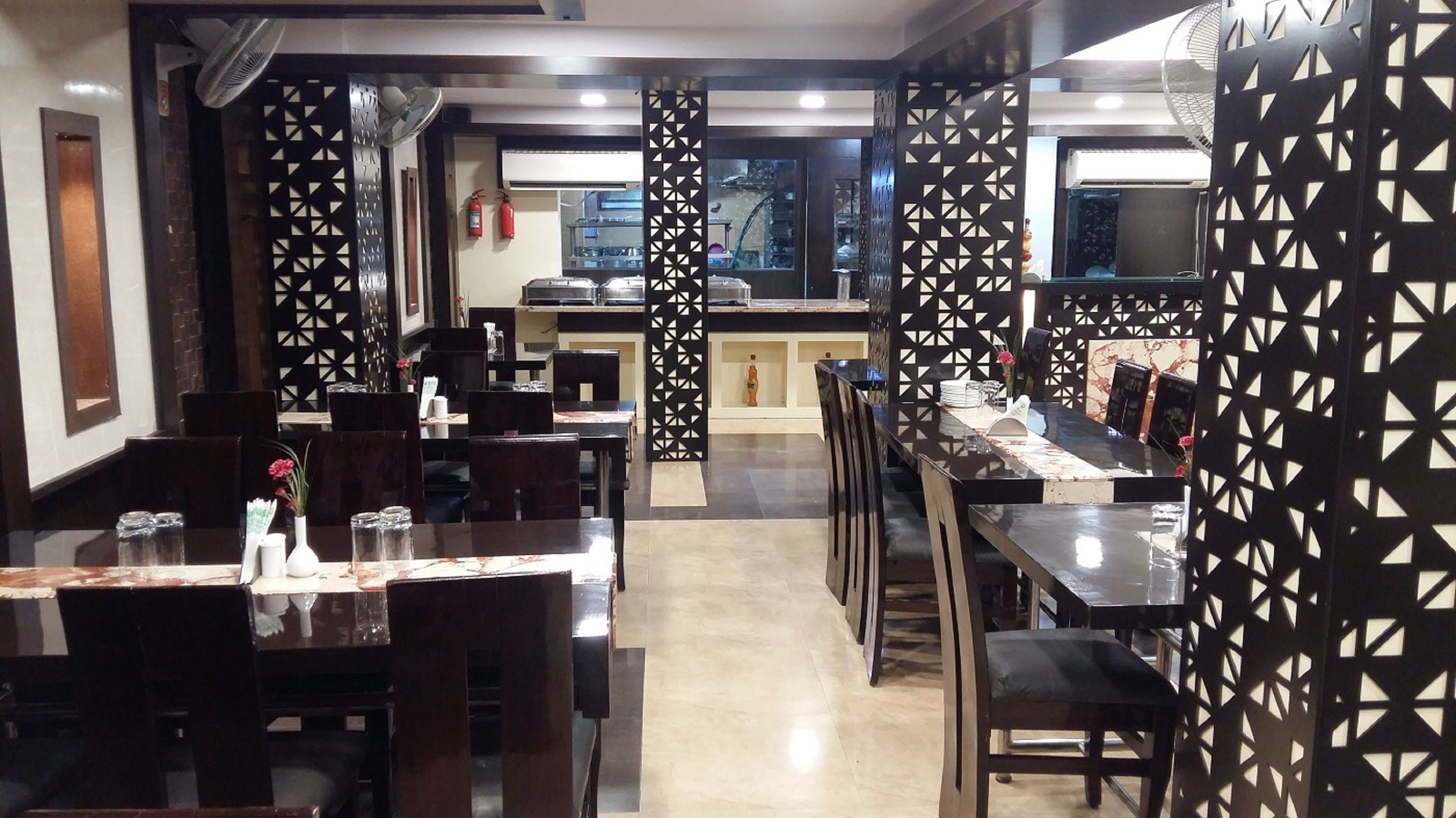 Un restaurante con mesas y sillas de madera oscura, paredes con diseños geométricos y una cocina visible al fondo.