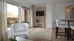 Una sala de estar moderna con un sofá, una mesa de comedor y sillas. Hay un televisor montado en la pared, una mesita con una planta y una ventana con cortinas.