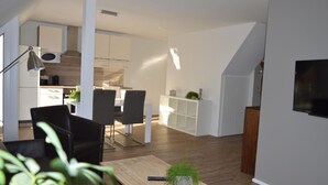 Una sala de estar moderna con piso de madera, un comedor con sillas negras, una cocina con gabinetes blancos y un televisor montado en la pared.