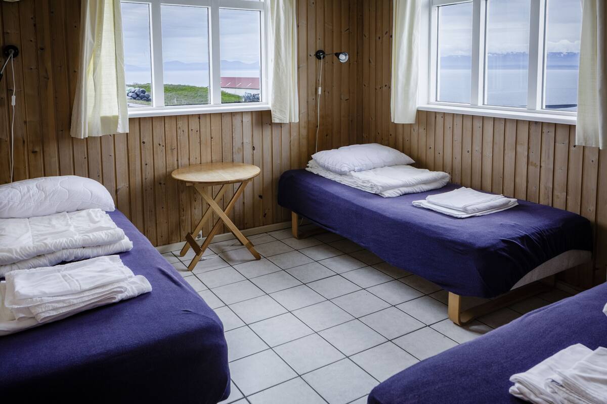 A room with two beds, a small wooden stool, and a window with a view of the outdoors.