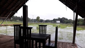 A wooden table and chairs under a thatched roof, overlooking a rice paddy field. - Kamatha Resort (Polonnaruwa)