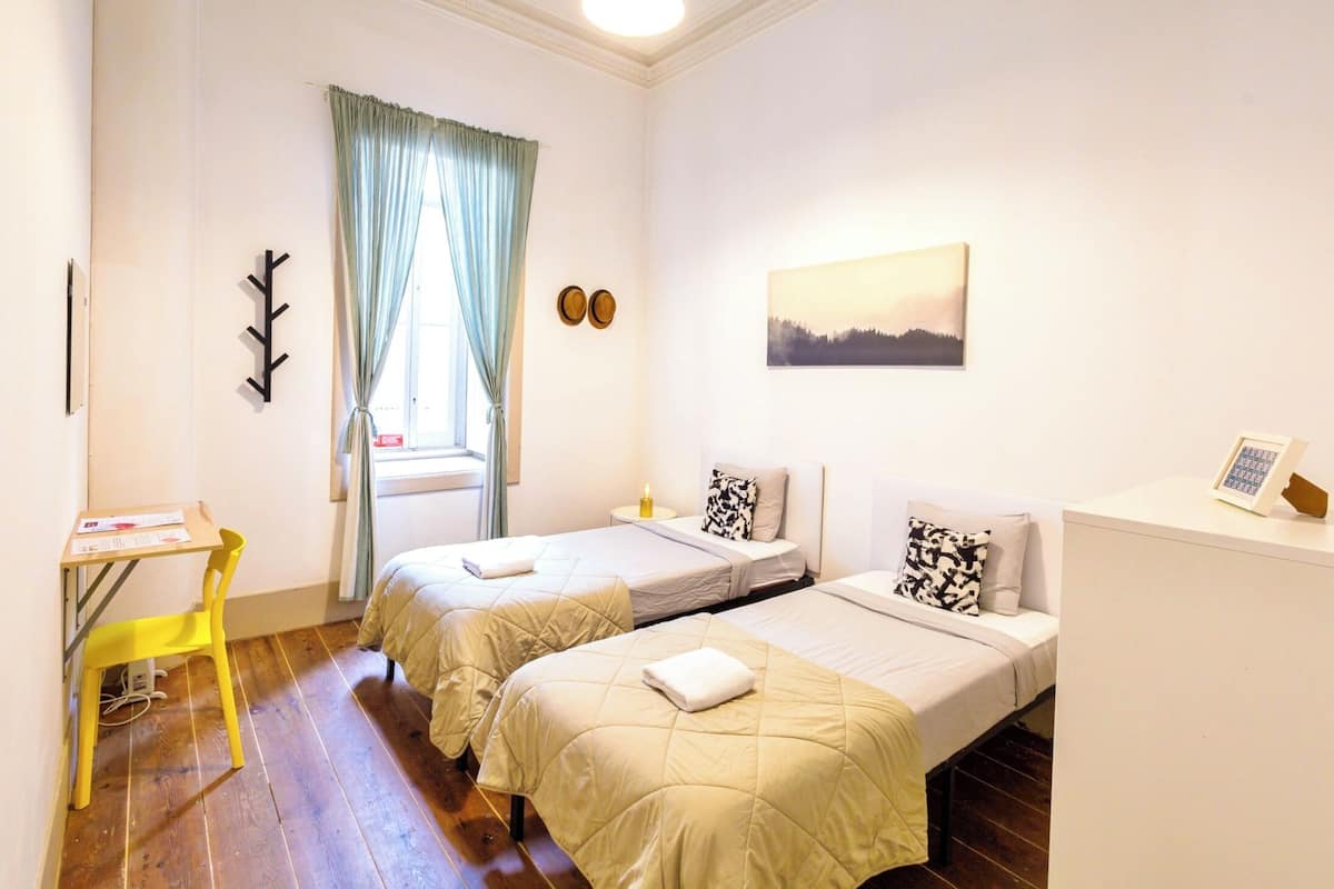 Une chambre avec deux lits, un bureau, une chaise, une fenêtre avec des rideaux, un porte-manteau fixé au mur et un tableau encadré.