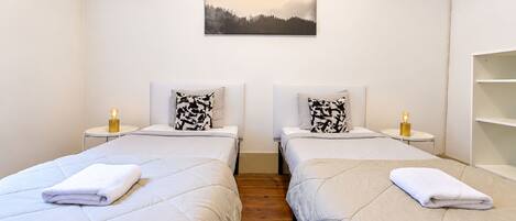 A hotel room with two beds, a bedside table with a lamp, a wall-mounted picture, and a wooden floor.