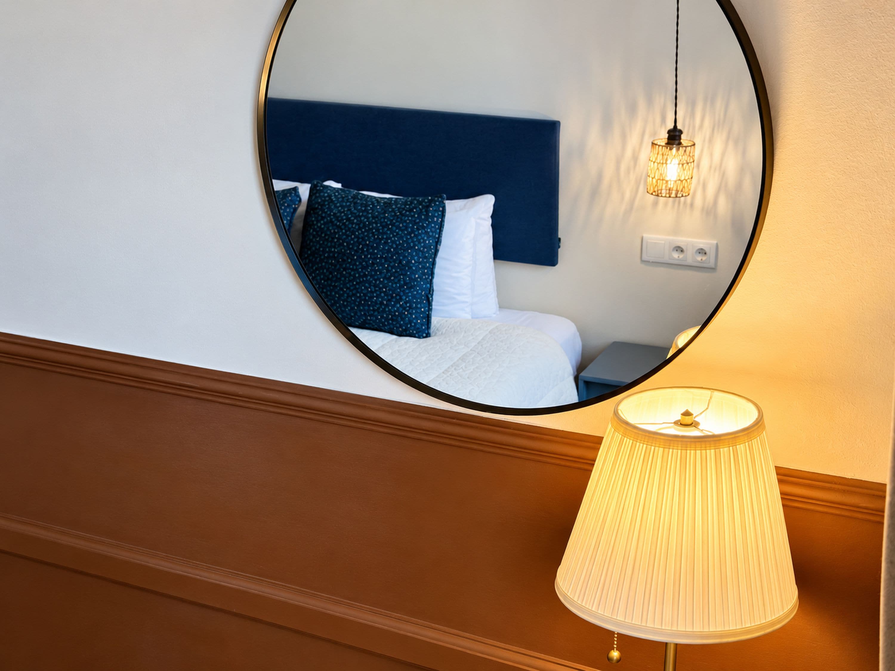 Ein Hotelzimmer mit einem runden Spiegel, der ein Bett, Kissen und einen Nachttisch reflektiert. Ein Lampenschirm ist auf dem Boden sichtbar.