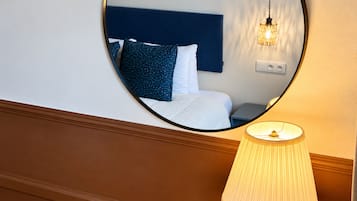 Ein Hotelzimmer mit einem runden Spiegel, der ein Bett, Kissen und einen Nachttisch reflektiert. Ein Lampenschirm ist auf dem Boden sichtbar.