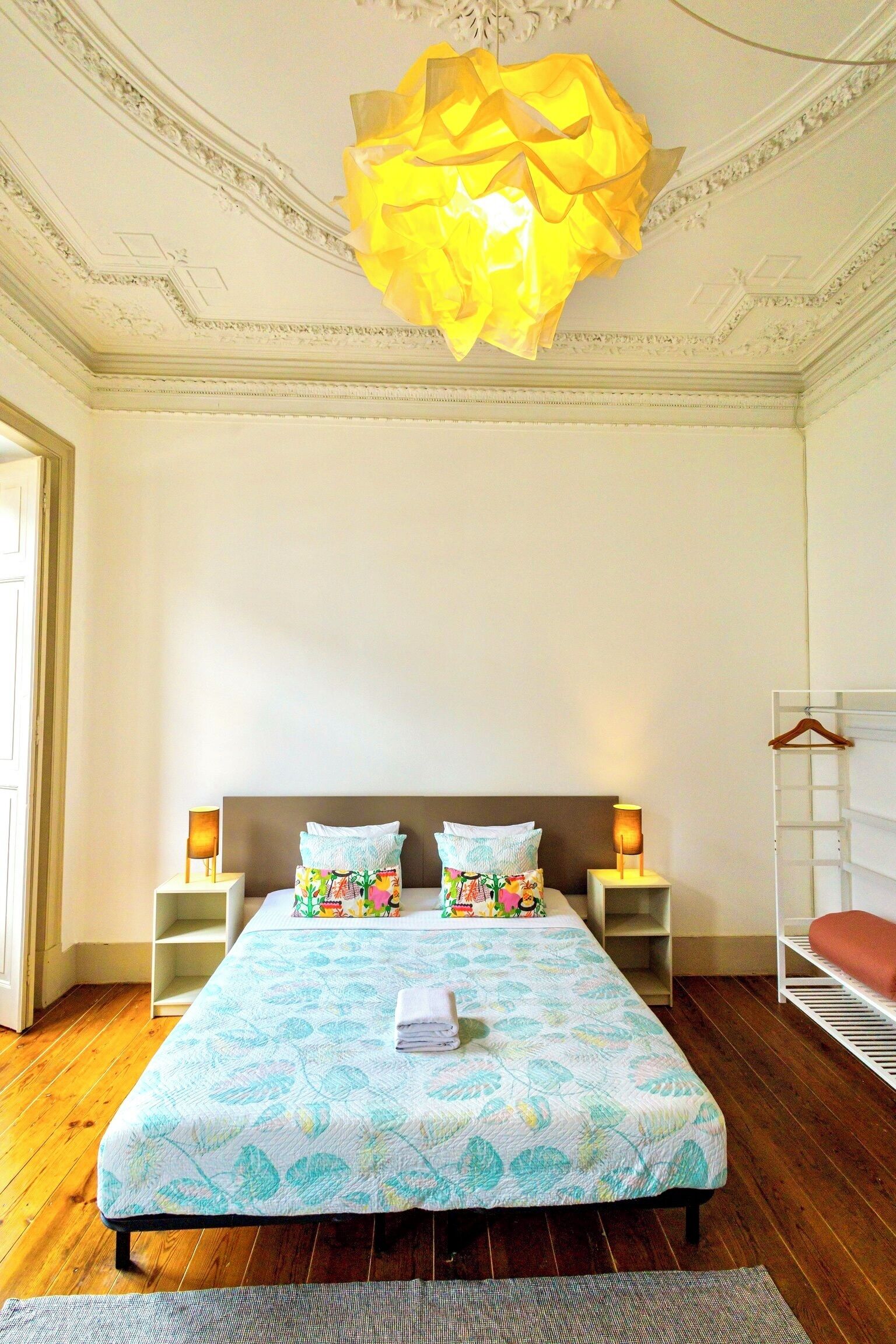 A bedroom with a large bed, two bedside tables with lamps, a hanging light fixture, and a wooden floor.