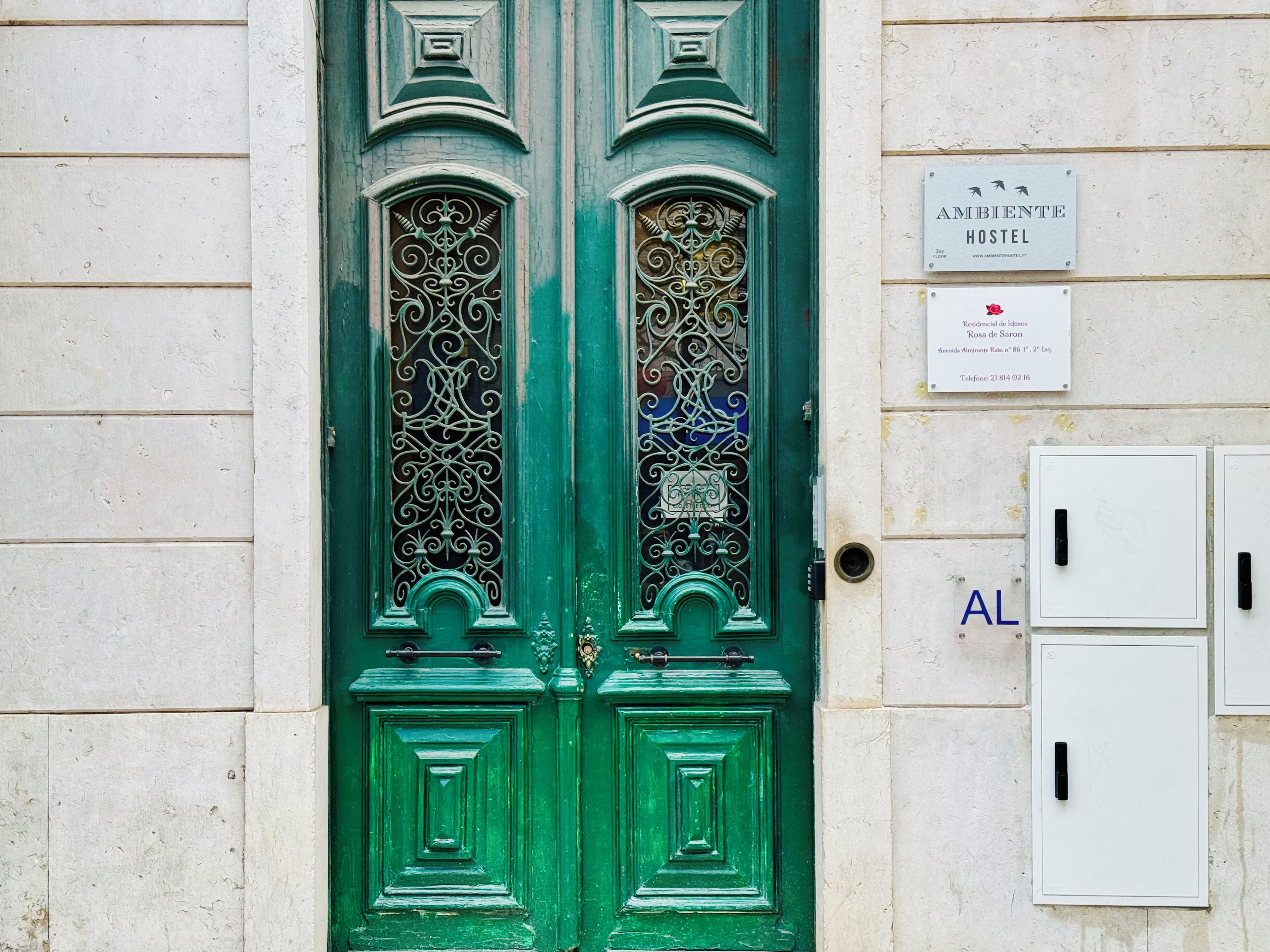 A green door with ornate metalwork, a sign for 'AMBIENTE HOSTEL', and a utility box with a black handle.