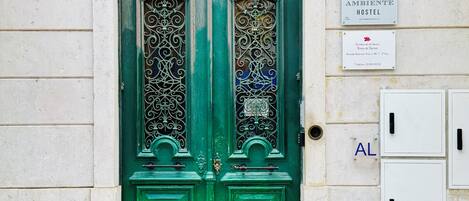 A green door with ornate metalwork, a sign for 'AMBIENTE HOSTEL', and a utility box with a black handle.