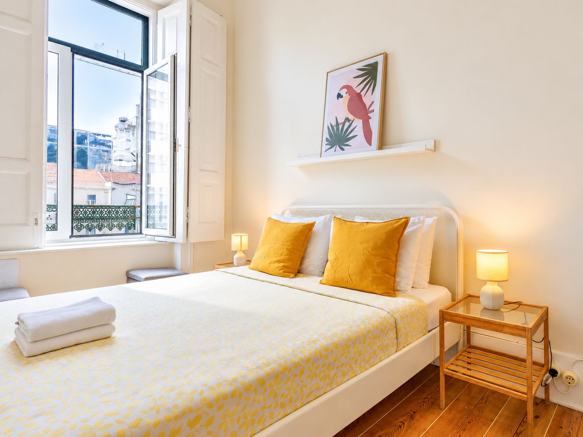Habitación de hotel con cama, mesita de noche, lámpara, ventana con vista a edificios y un cuadro colgado en la pared.