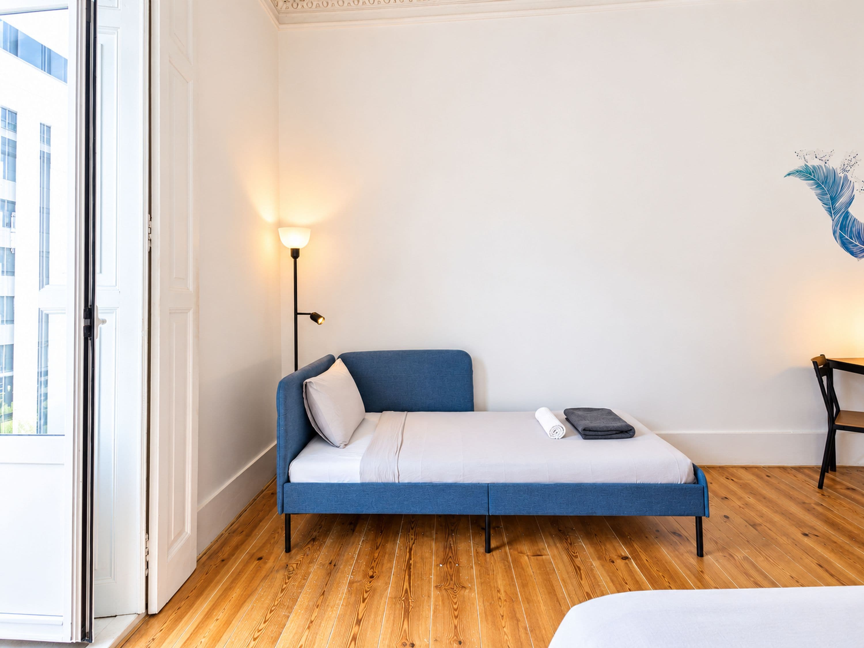 A room with a blue sofa bed, a bedside table with a lamp, and a view of the outside.