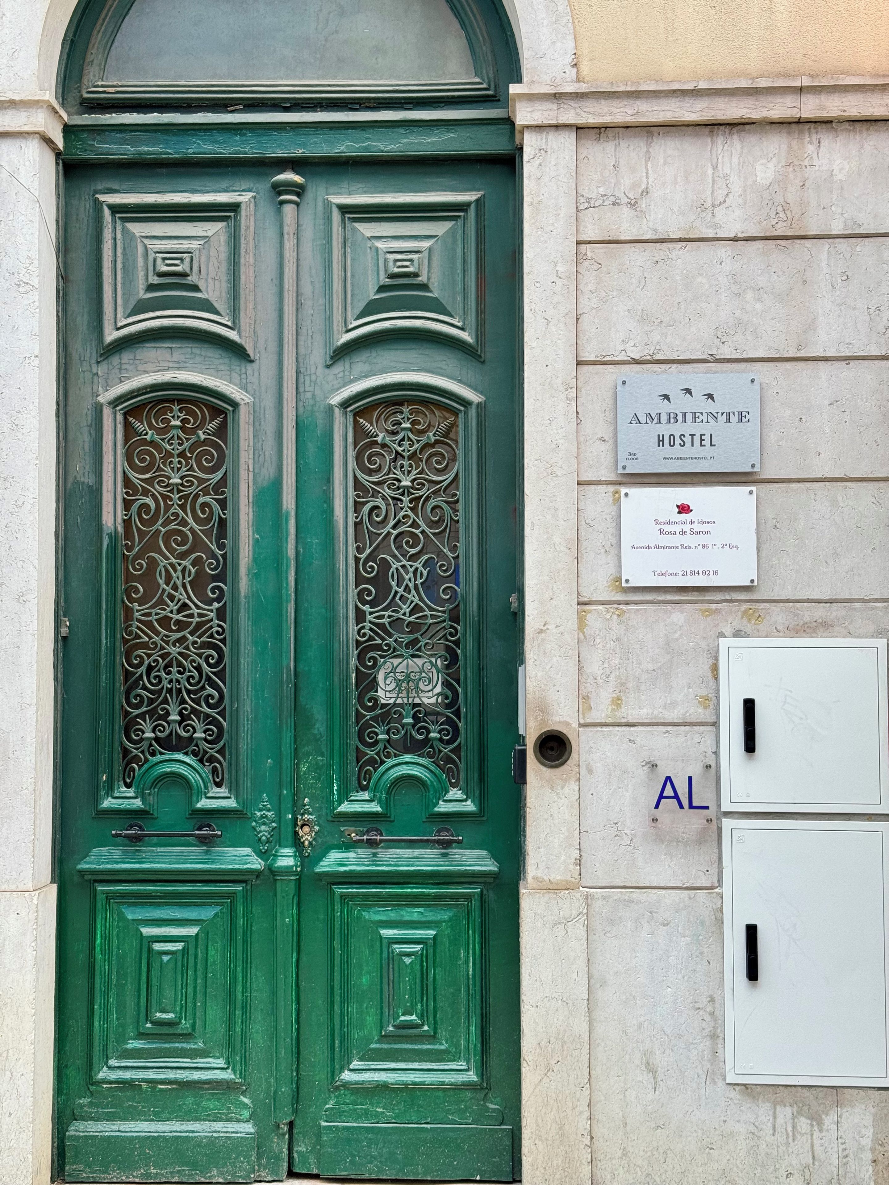 Une porte verte à deux battants ornée de motifs métalliques complexes, une enseigne indiquant « Ambiente Hostel » sur le mur, et un petit panneau indiquant « Rosa de Saron ».